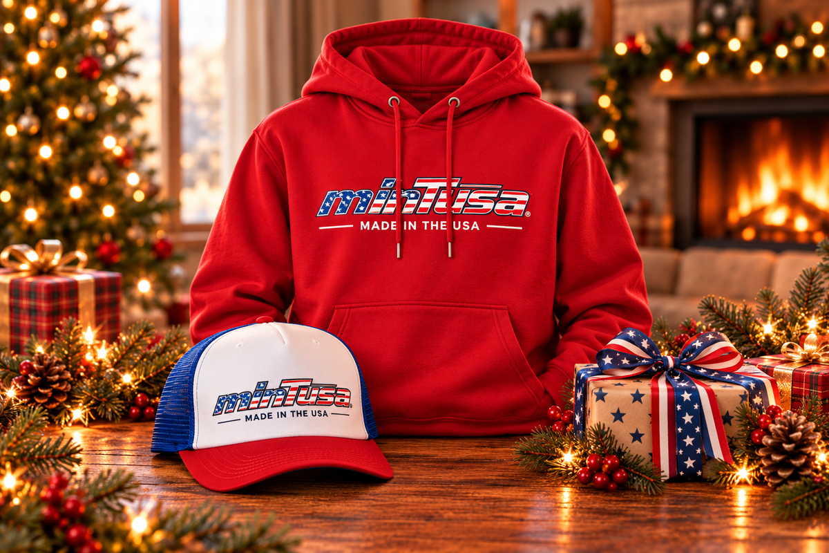 minTusa Star Spangled Holiday Set: a red hoodie and a red, white and royal foam trucker hat, both with the flag-pattern minTusa logo, beside a star-wrapped gift box by a Christmas tree and fireplace