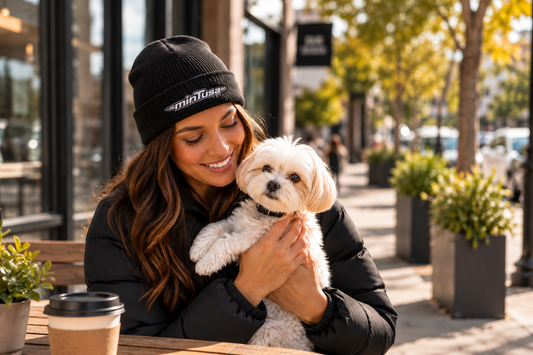 Smiling woman holding her small white dog at a sidewalk cafe, wearing a black minTusa Classic fisherman beanie